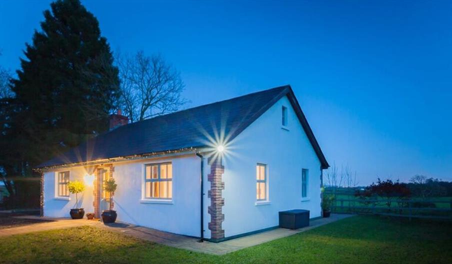 A cottage at night beside a country feild in front of a large tree.