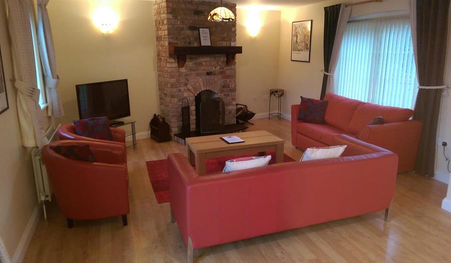 Image shows lounge area with sofas, chairs, TV and fireplace. Two windows in the room.
