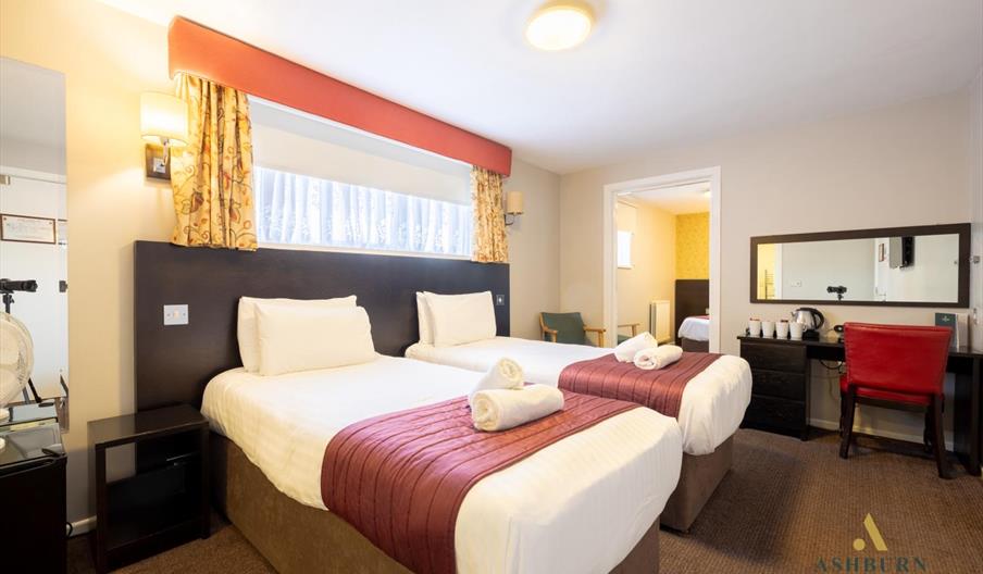 Bedroom with two single beds, white bedding, red runners, a vanity desk with a mirror, and an open doorway leading to a bathroom.