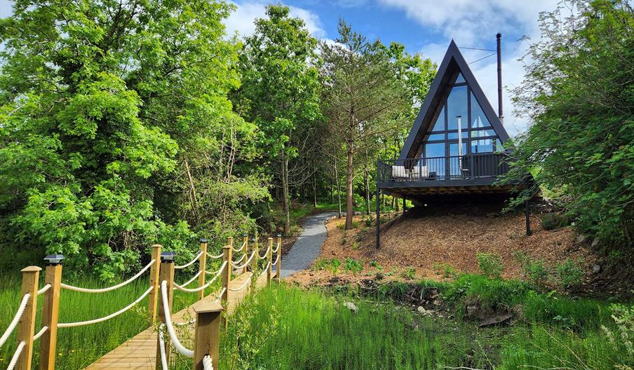 A luxury A-frame lodge set in peaceful woodland surroundings, overlooking a pond with a wooden rope bridge leading through the trees.
