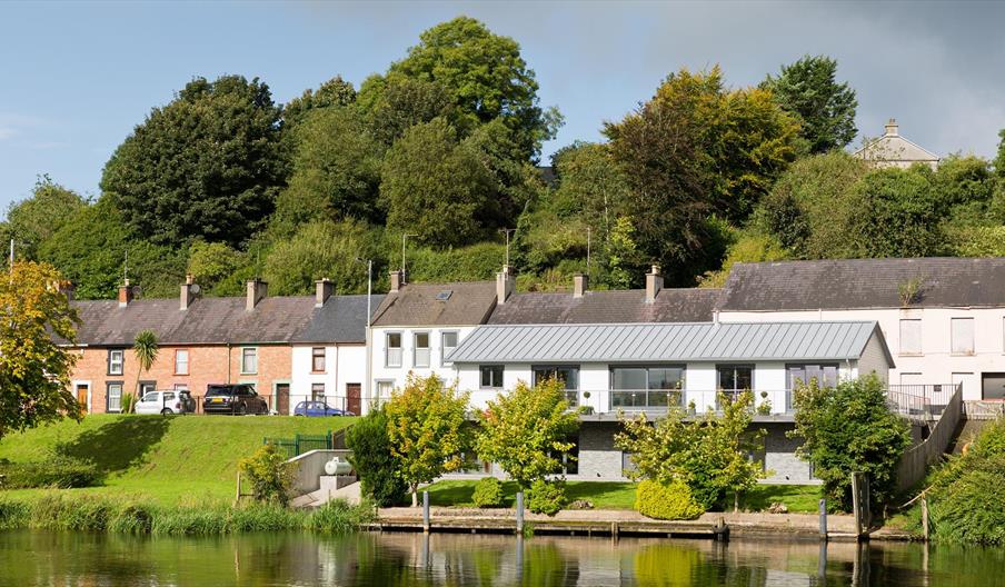 Riverside view of River Cottage with large windows and balcony, set along the water’s edge with mature trees and traditional terraced houses behind.