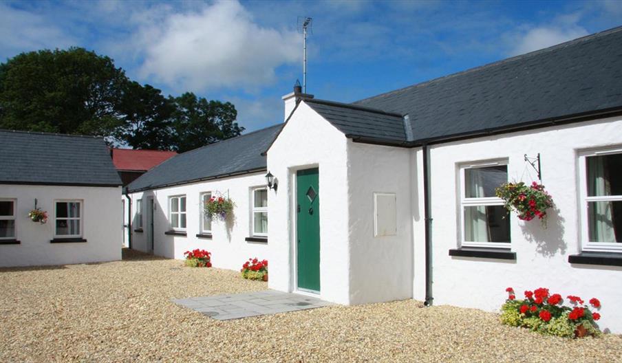 Image shows front of whitewashed property with green door and gravel drive. Flowerbeds on gravel outside windows