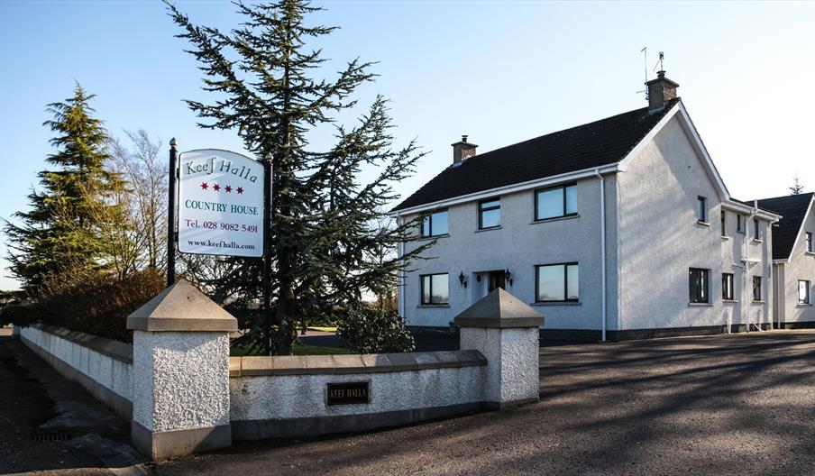 A roadside view of a large country house with a sign and trees outside.