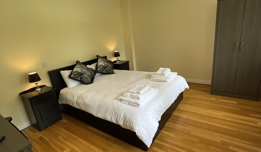 Double bedroom with hard floor wooden floor. Double bed dressed in white linen, bedside cabinets on both sides of the bed. Wardrobe and side of dressi