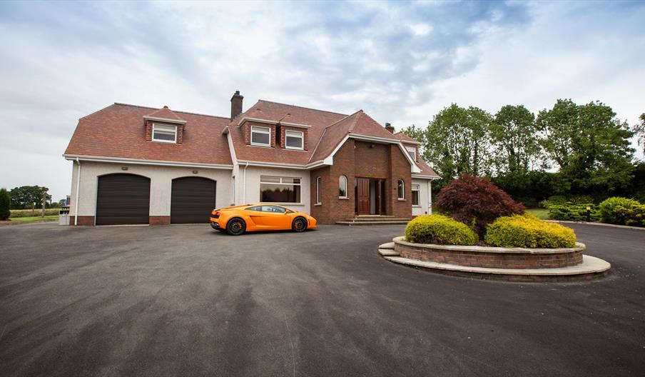 Image shows front of property with orange coloured super car in the large driveway