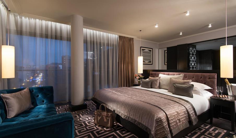 A large double bedroom with a blue sofa, champagne in an ice bucket and a city view at night.