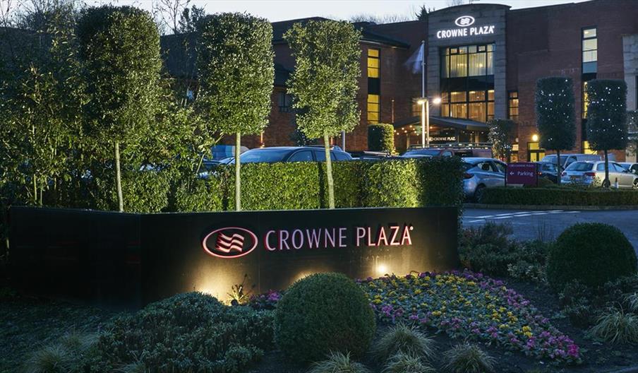 Image of the outside of the Crowne Plaza Belfast hotel. Flower bed with "Crowne Plaza" sign in the foreground, hotel and parking lot in the background
