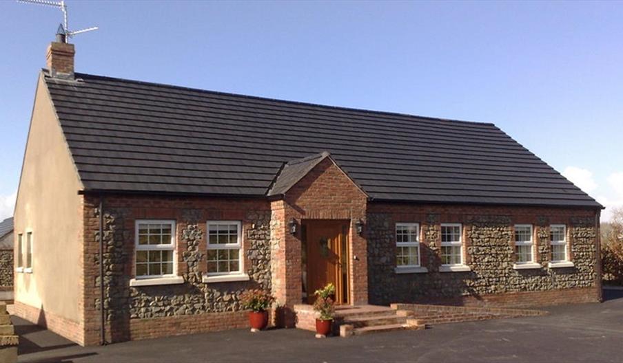 Image shows front of detached cottage which has 6 windows and front door with a few low steps