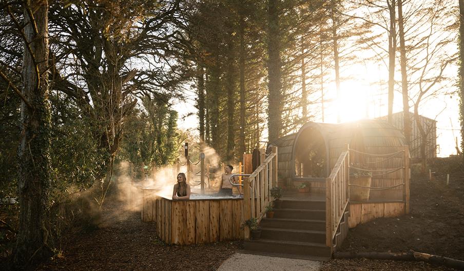 A couple perched on the edge of a hot tub elevated above the Forest floor. Views behind them of the River Maine.