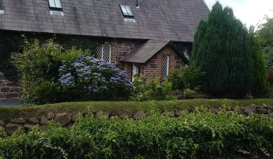 Image is of front of cottage with stone wall covered with plants and flowerbeds