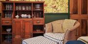 A sitting room with a chair, footrest and a cupboard with glasses and a tea set.