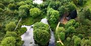 Aerial view of a luxury A-frame lodge nestled among woodland, with ponds, walking paths and green countryside surrounding the retreat.