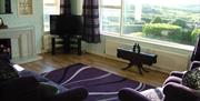 Image shows lounge with TV, wooden floor, rug, sofa and armchair looking out to view of garden and beyond