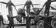 Black and white photograph showing acrobatic circus performers in front of a circus tent and cart - Three man stand on the base with three other men s