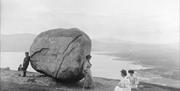 Black and white photograph showing a group of two men and three women sitting beside a large boulder on a hill, overlooking a bay and town in the dist