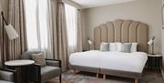 Contemporary hotel bedroom with king-size bed, cushioned headboard, seating area and large windows with natural light.