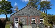 Image shows stone cottage covered in ivy at the right hand side plus white front door and bench outside