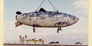 A mosaic sculpture in the shape of a fish is suspended by a crane while being installed on Donegall Quay. In the background, the bright yellow Harland